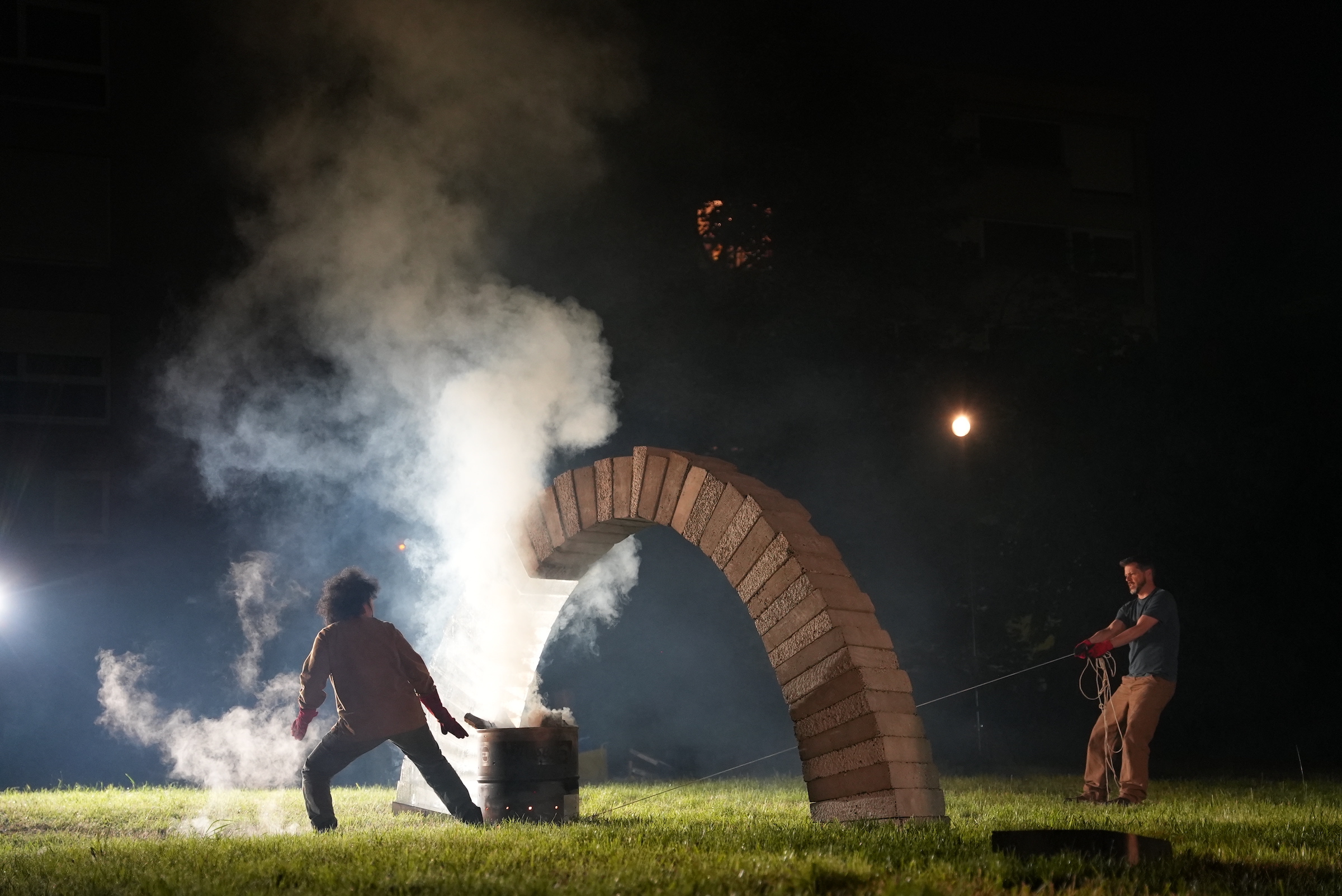 Outdoors at night. Two people drag and push a fire burning in an oil drum under a freestanding arch, about 2 metres tall and 4 metres wide, made out of concrete and ice blocks. The fire is smoking and the people are silhouetted against it.