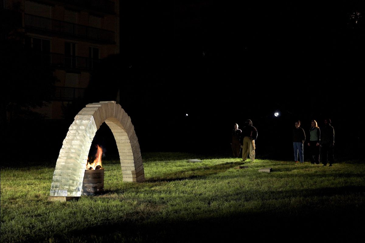 Outdoors at night, an arch made two thirds from concrete blocks and one third of ice blocks stands over a fire, which burns in an oil drum below it. The arch is lit by a pool of electric light, on the edge of the light, 6 singers stand watching and singing.  