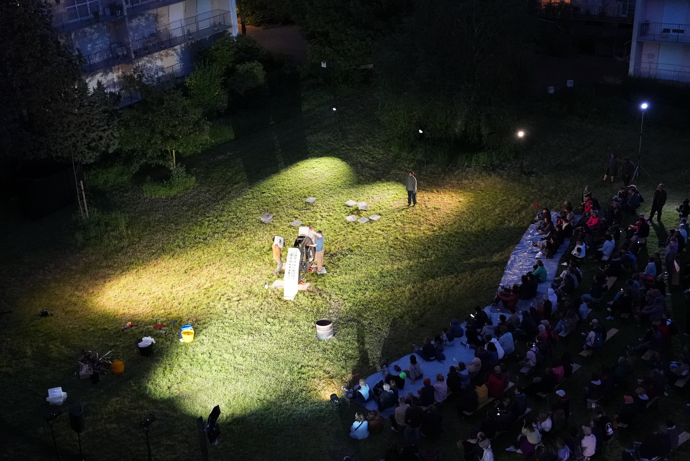 A grassy courtyard at night surrounded by apartment buildings seen from above. In the centre of the space lights point towards an arch being built by two people, as one person sings and watches them. Rows and rows of audience are seated and standing around, watching them.