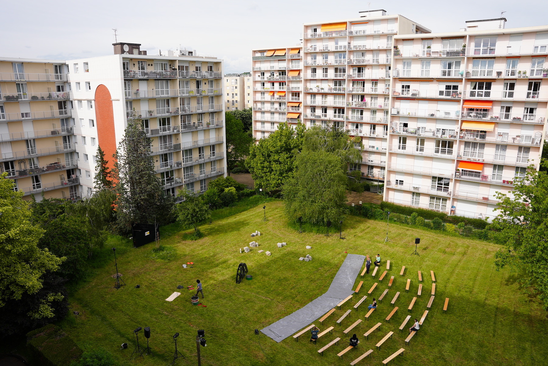 Daytime, outdoors. A grassy courtyard surrounded by apartment buildings seen from above. In the centre of the space lights and prop pieces are set up before show time. Rows of seating await an audience.