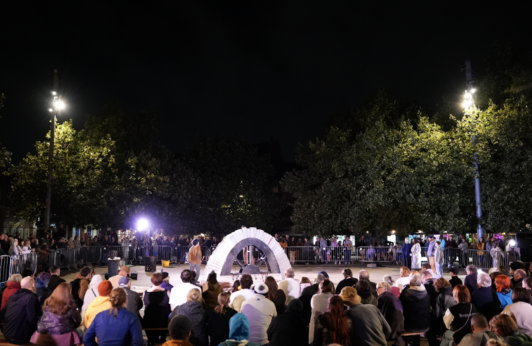 Outside at night. An audience surrounds a performance space, bathed in light. An arch is complete, supported by a wooden former. Performers stand singing to it. 