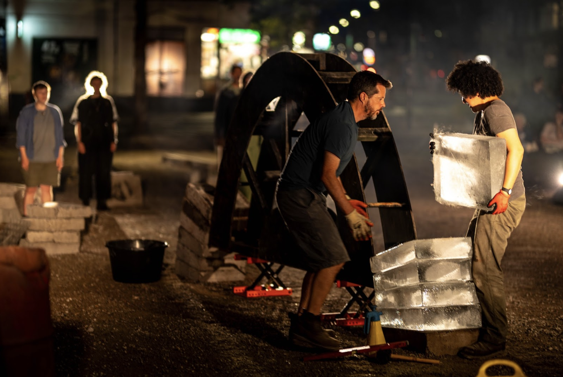 Outside at night. Two performers lay ice blocks on top of each other in the early stages of the construction of an arch. In the background performers stand and sing. 