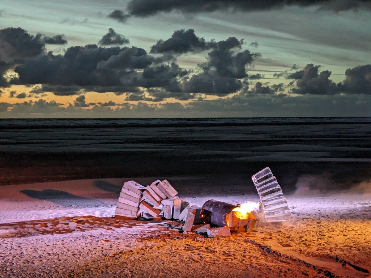 Outdoors on a beach. The collapsed arch and fallen oil barrel with the fire still burning sit in front of the sea and dusky sky. Smoke and long shadows add to the mood.