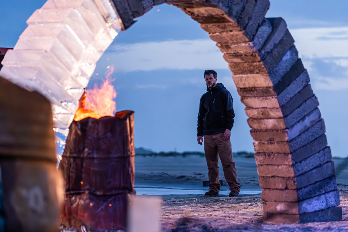 Outdoors on a beach in the dawn light. A performer is seen standing framed by the arch of concrete and ice. A fire burns brightly in an oil barrel beneath it.