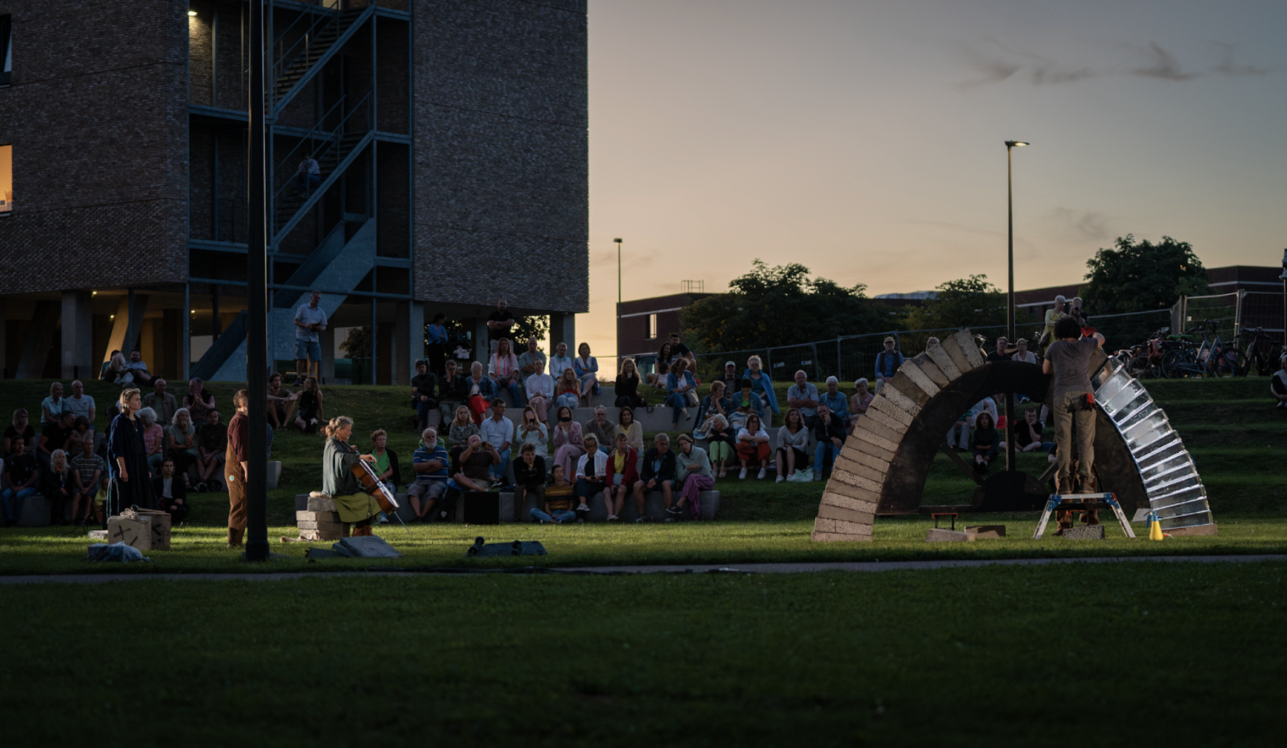 Outside at dusk, to the right the arch is nearly completely constructed. A performer stands in front of it on a low step to reach the higher part of the arch. To the left three performers, two standing and one seated with a cello, look towards the arch, singing and playing. Beyond them a bank of audience look on, underneath a darkening clear sky.  