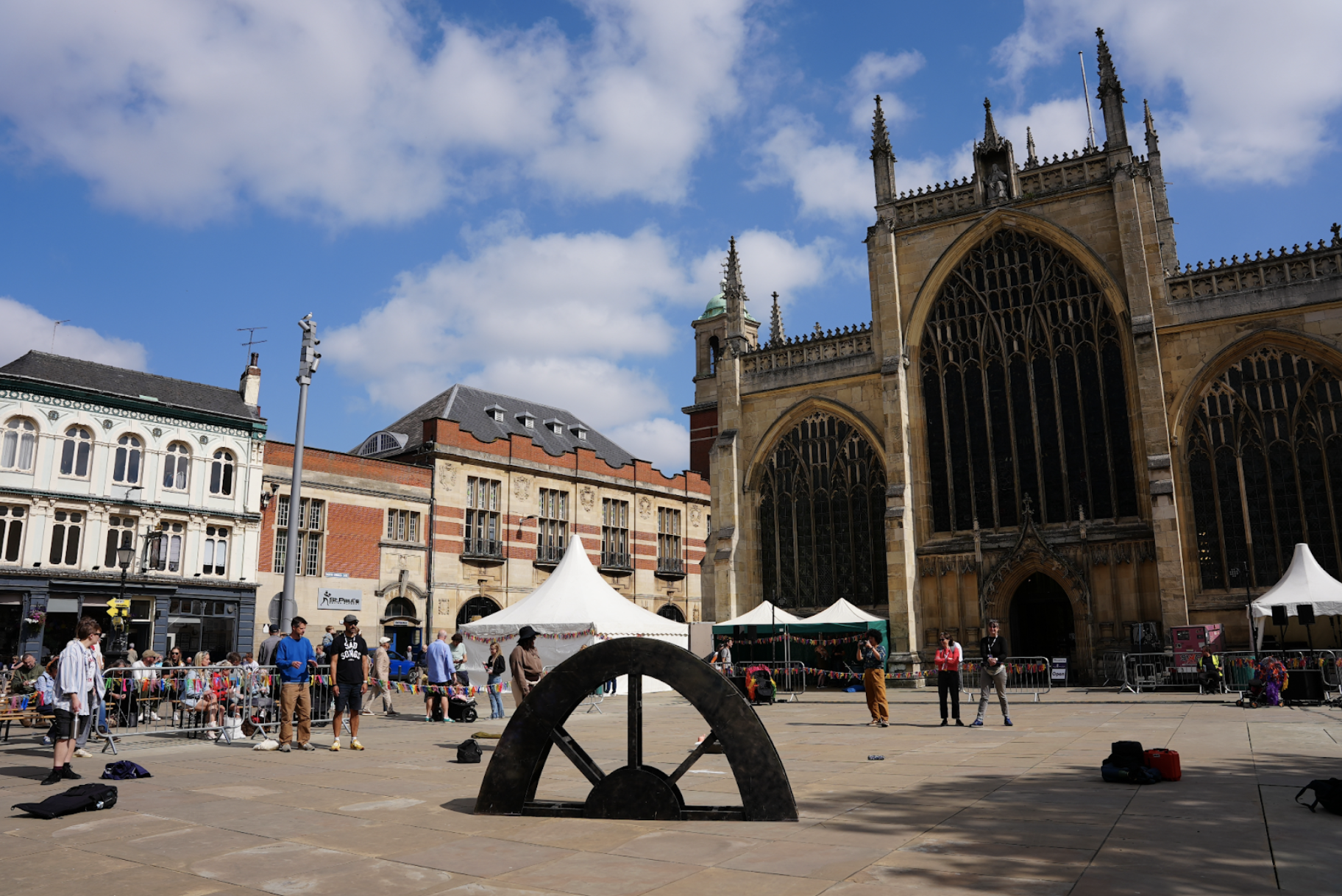 Daytime outside. An urban square is surrounded by buildings including an ornate minster with three large arched windows. In the foreground a wooden arch former stands. around the former performers rehearse, standing. Beyond them, some seated public watch the rehearsal. 