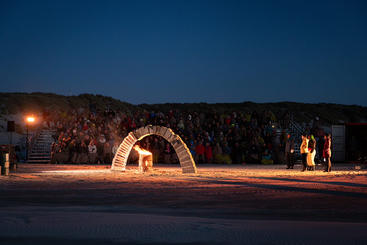 A beach, outdoors at night. A fire is burning under a freestanding arch made two thirds of concrete blocks and one third of ice blocks. An audience of hundreds of people watch on, wrapped in blankets. Performers face the arch, singing.