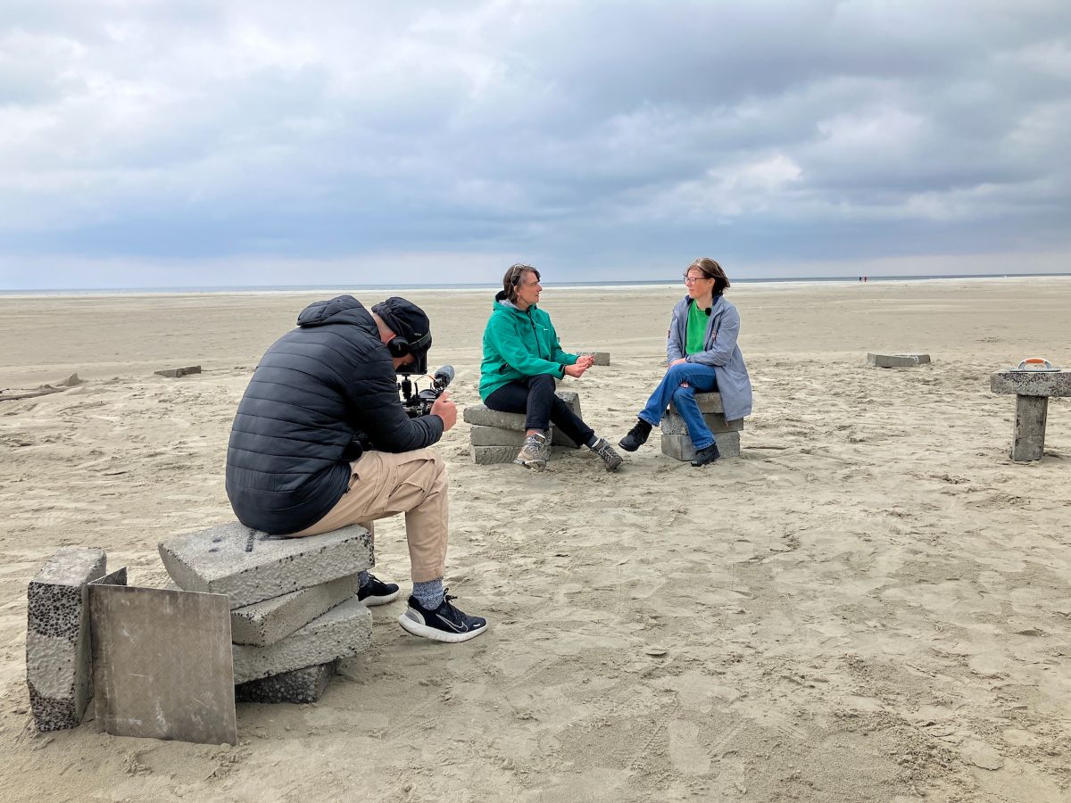 Outdoors on a vast beach. It’s a windy day. Two people sit on the set in conversation while a videographer films them talking.