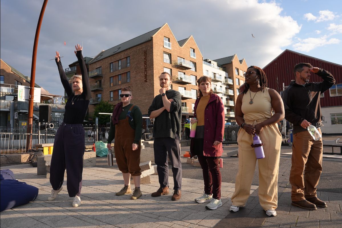 Daytime, outdoors. Six performers stand in the performance space as they prepare for the show in central Bristol.