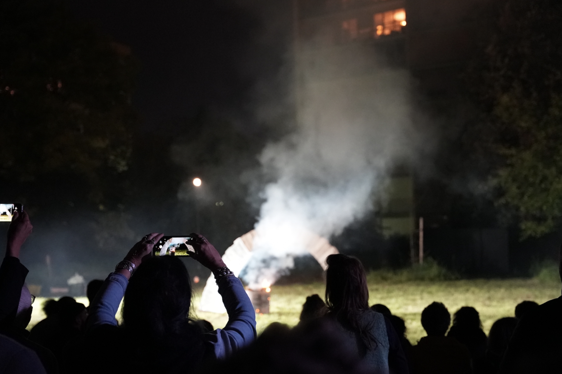 Audiences hold up mobile phones to take photographs. They are silhouetted against a bright arch on green grass, with a fire under it, smoke rising. This arch is the subject of their photographs, apparent on the lit screens of their phones. . 