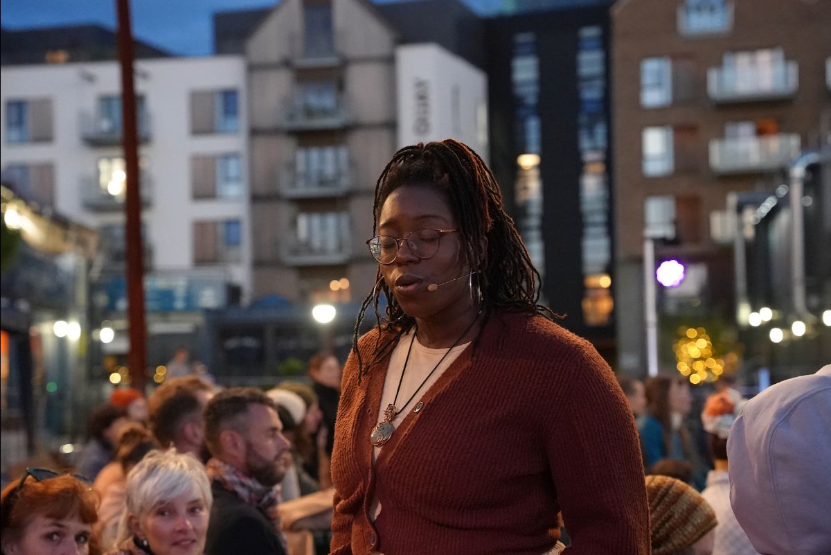 Evening, outdoors. A performer in arch, stands among the audience and sings, her eyes closed and her eyebrows raised in concentration.