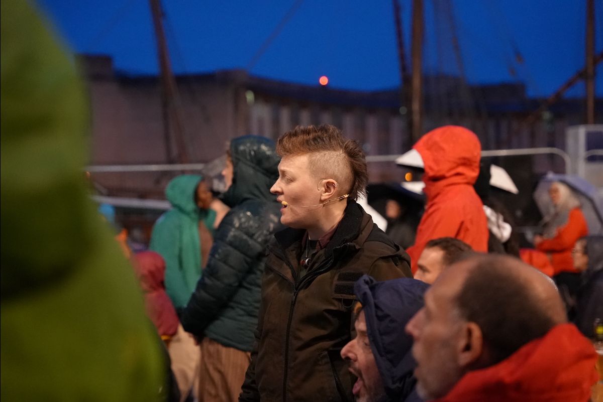 Evening, outdoors. One of the performers in Arch, stands amongst the audience, singing.