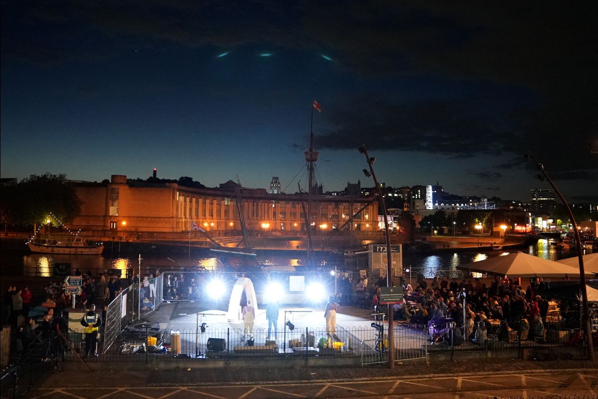 Night-time, outdoors. A fenced in performance of Arch in Bristol city centre. Audience are watching both inside and outside the fence. The harbourside, buildings and night city lights surround the scene.