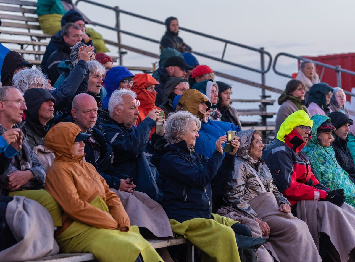 Evening, outdoors. A dedicated crowd wrapped in blankets and waterproofs watch intently.