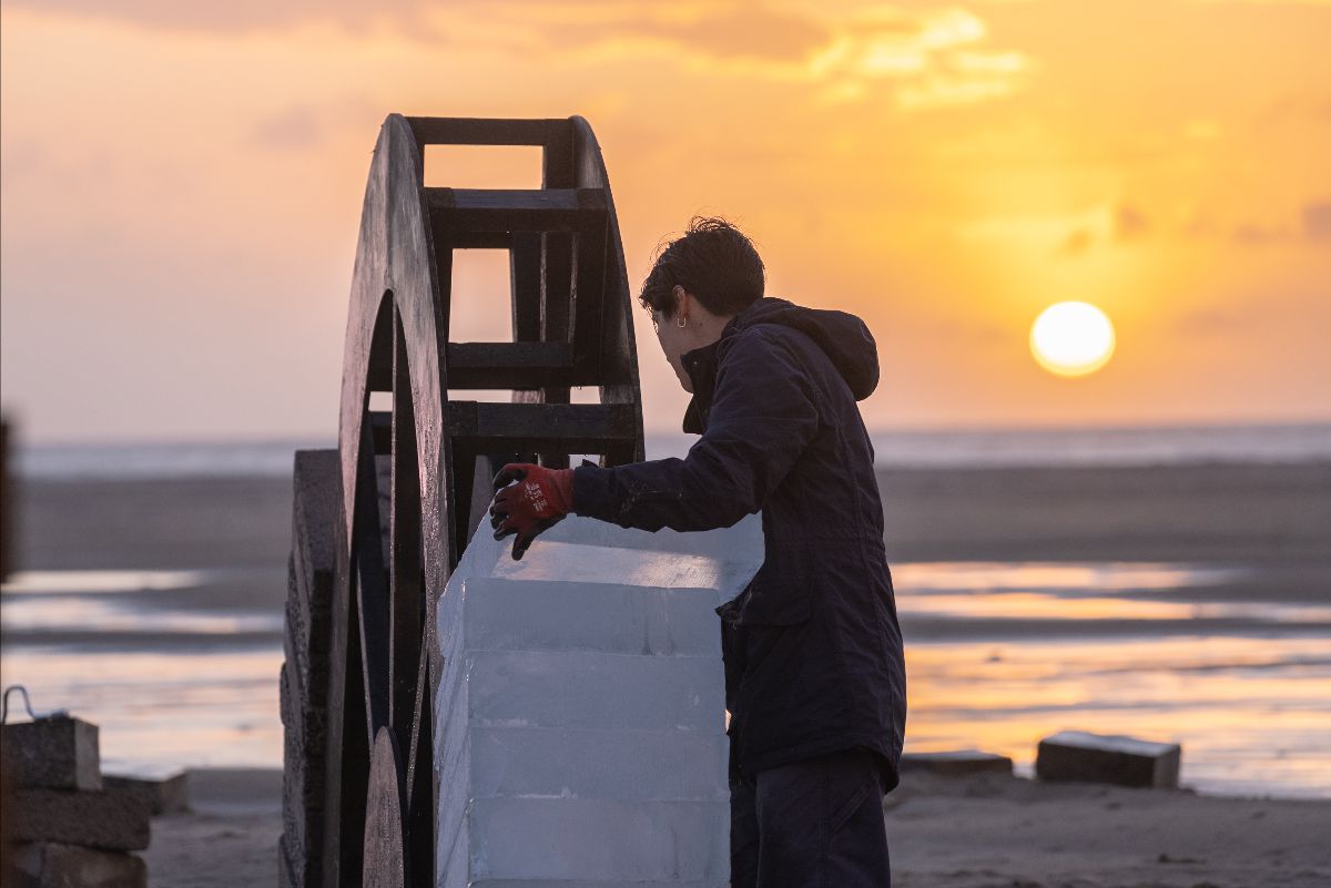 Outdoors on a flooded beach. The setting sun is almost at the horizon where the sea meets the sky. A performer places an ice block on the existing stack around the former.