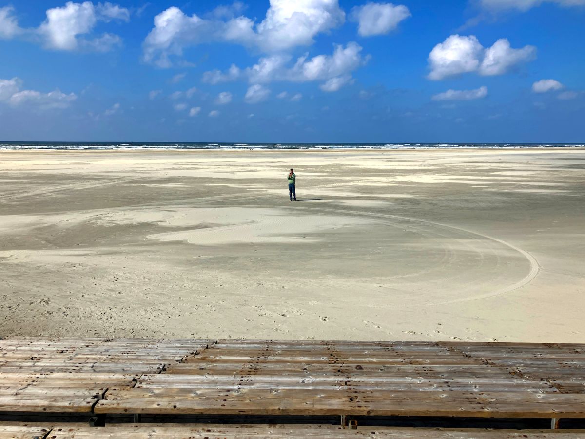 Daytime on a vast beach. A sole figure stands on an empty beach beyond empty wooden bleachers. Sea and blue sky on the horizon.