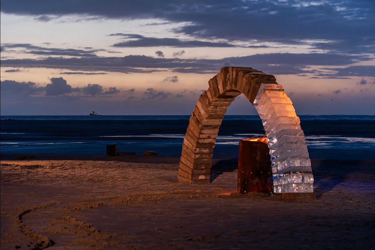  A beach, outdoors at night. A fire is burning under a freestanding arch made two thirds out of concrete blocks and on third of ice blocks. The sea and wispy clouds in the pink sky form the backdrop.