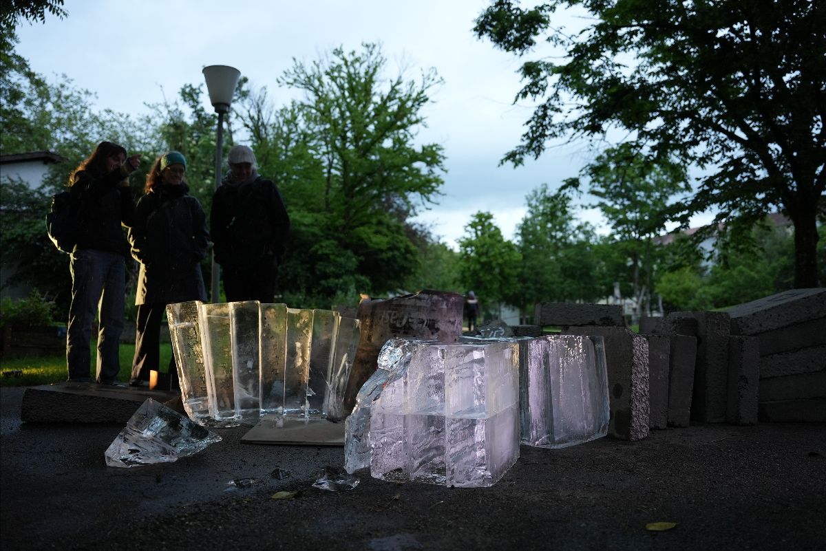 Ice and concrete blocks lie strewn on the ground outdoors at dusk. Three people stand behind them, talking and pointing, silhouetted against the fading evening light.