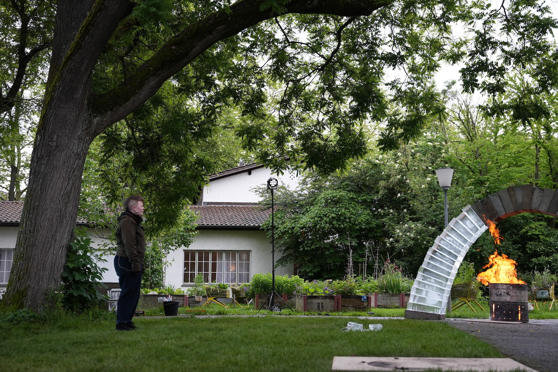 A grassy outdoor space surrounded by trees. The arch, made out of concrete and ice blocks, stands over a roaring fire. To the left, a performer is singing to the arch.