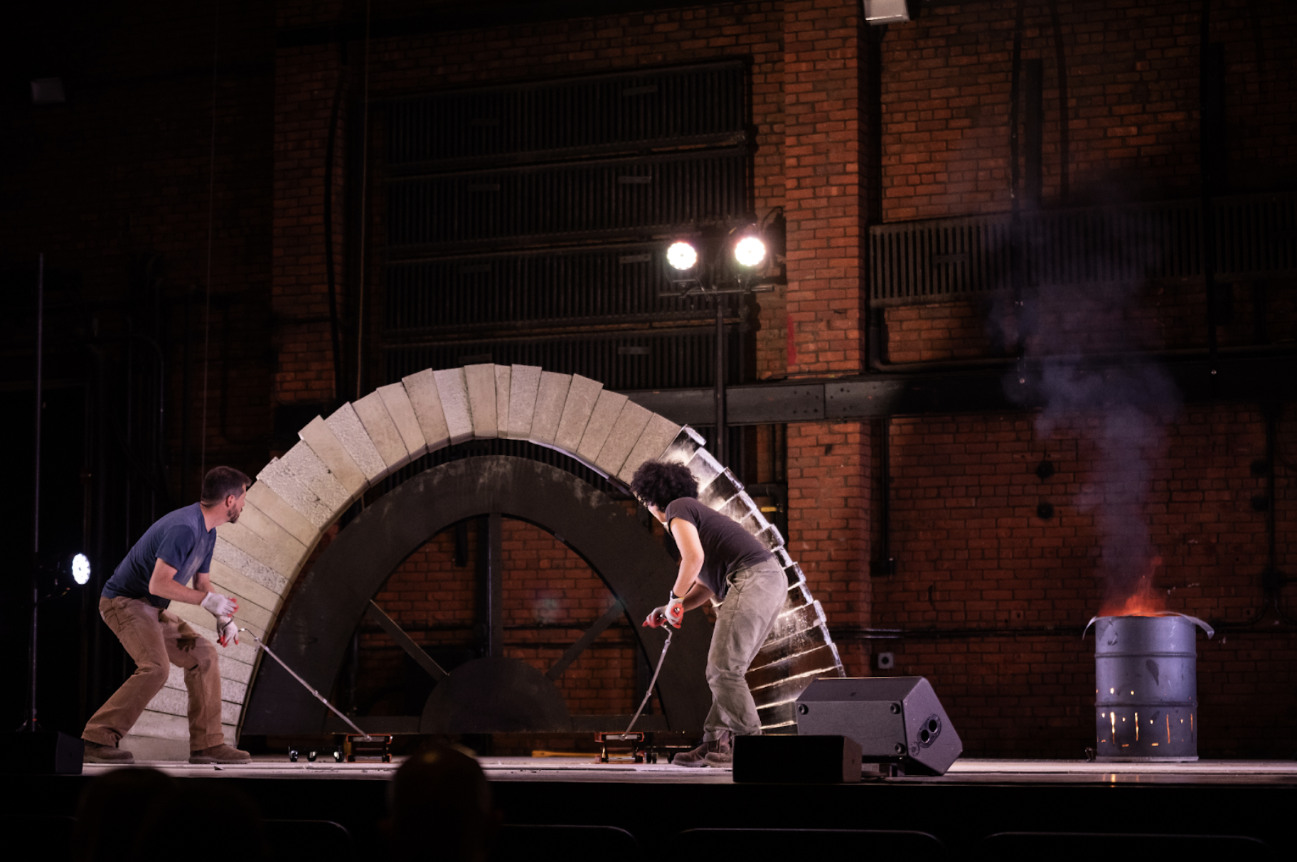 A stage, lit by electric light. Two performers, attentive to their task, remove a wooden former under an arch made two thirds of concrete blocks and one third of ice blocks. A fire burns in an oil barrel to the right of them. 