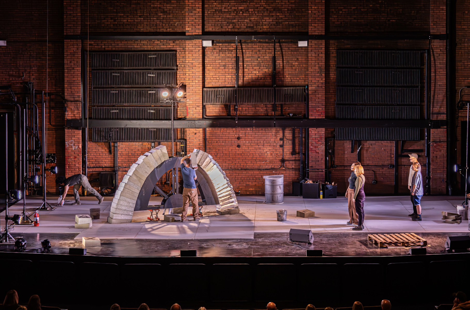 6 performers perform on a large stage, building a freestanding arch and singing. They are lit by the lights of the theatre, which casts shadows across the stage and onto a large brick wall of the back of the theatre. In the foreground are the first rows of audience watching from the auditorium. 