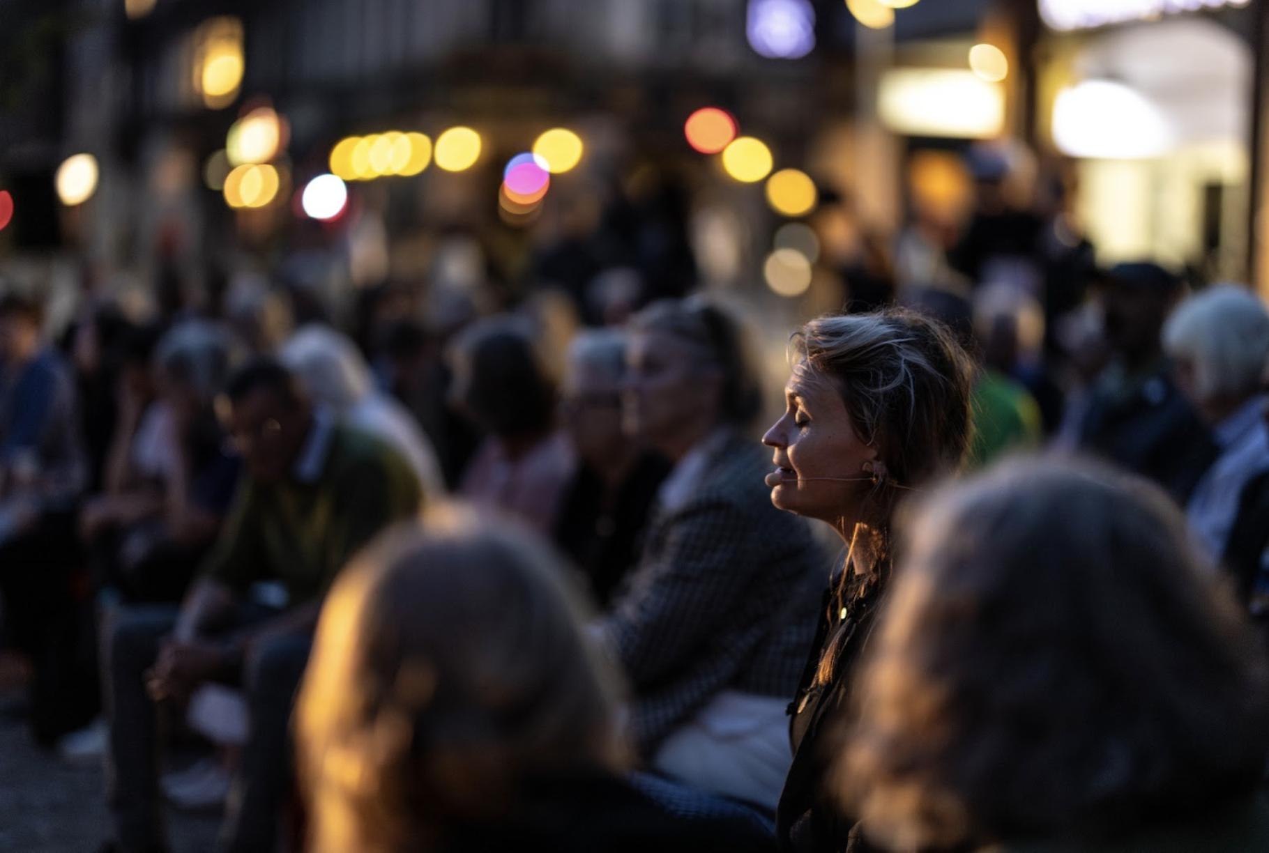 From amongst a watching audience, a performer sings, the light catching the contours of their face, their eyes closed and eyebrows raised in expressive concentration.  