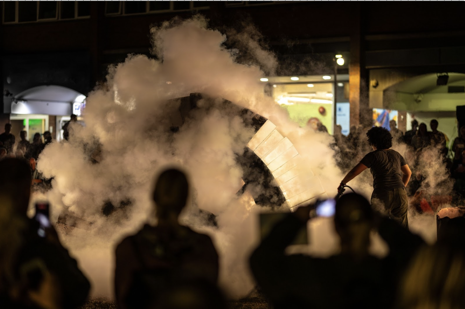 Outside at night. In the foreground audience are silhouetted, some holding up phones, with lit screens, to photograph the performance. A performer fires a fire extinguisher at a completed arch of concrete and ice, creating clouds of vapour which catches the light. Beyond, audiences watch on, silhouetted against the lights of the city behind them. 