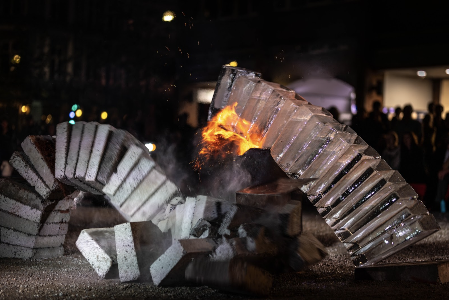 Outside at night. The arch of concrete and ice falls into the fire. In the foreground a curl of concrete blocks falls to the ground, hitting a barrel of fire at its base, which in turn is being crushed by a pillar of ice blocks falling onto it. In the background people watch, silhouetted against the lights of the city. 