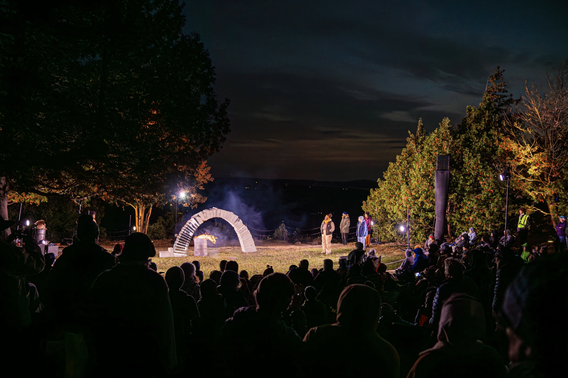 Outside at night, green grass surrounded by trees is lit with electric light. An audience watches 6 performers standing and watching a freestanding arch, made two thirds of concrete blocks and one third of ice blocks. An oil drum of fire burns under the arch. The sky has the last remaining light of the already set sun. 