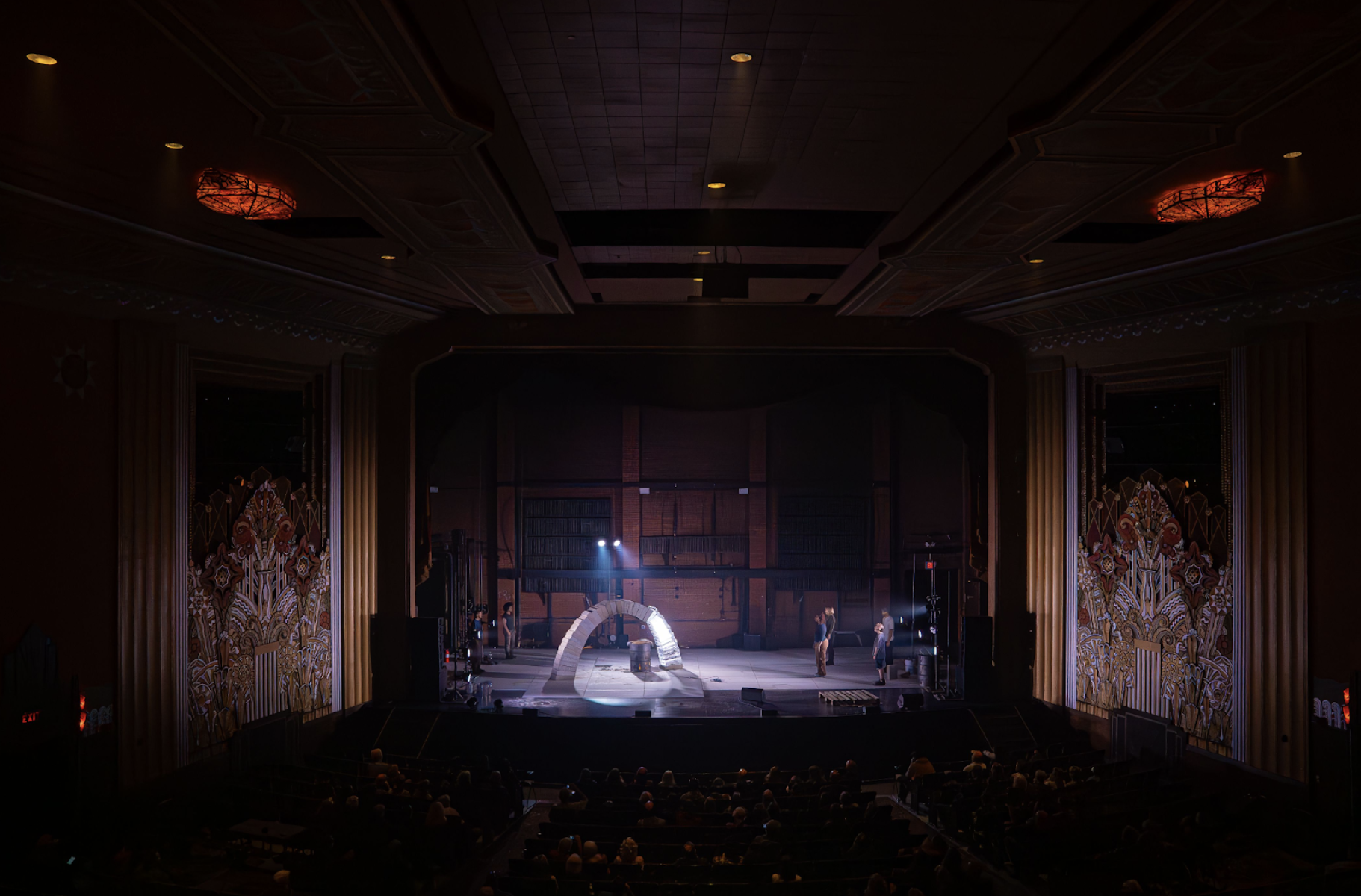 An auditorium with ornate walls, several rows of audience in the stalls, and, beyond, a stage. On stage 6 performers, lit in electric light, stand and watch as a fire burns under a freestanding arch made two thirds of concrete blocks and one third of ice blocks. Beams of light catch the smoke from the fire. 