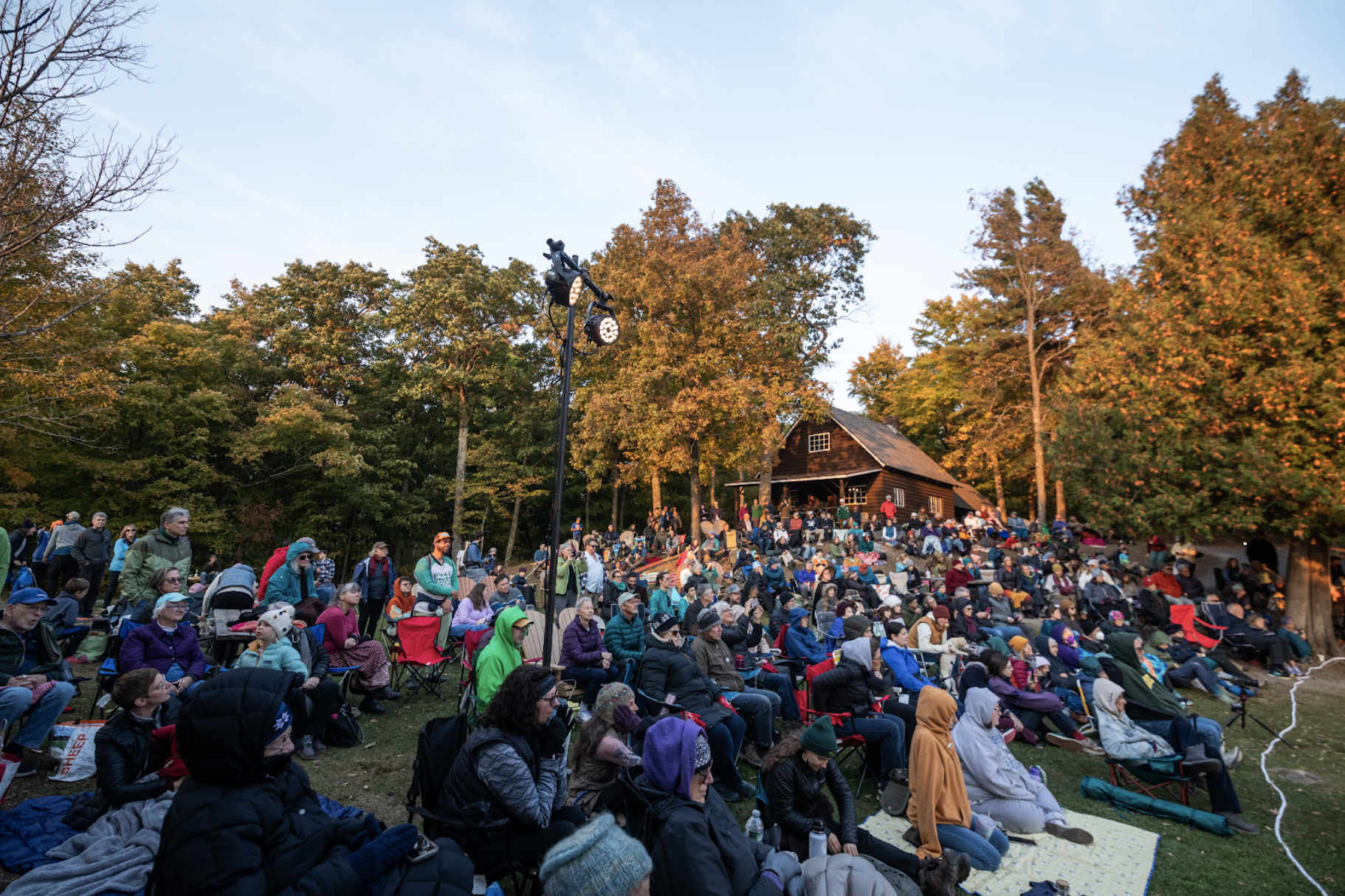 Many people sit on the ground on chairs and picnic blankets, all facing from left to right, half of them caught in the remains of the setting sun. Behind them autumnal trees catch the evening sun.