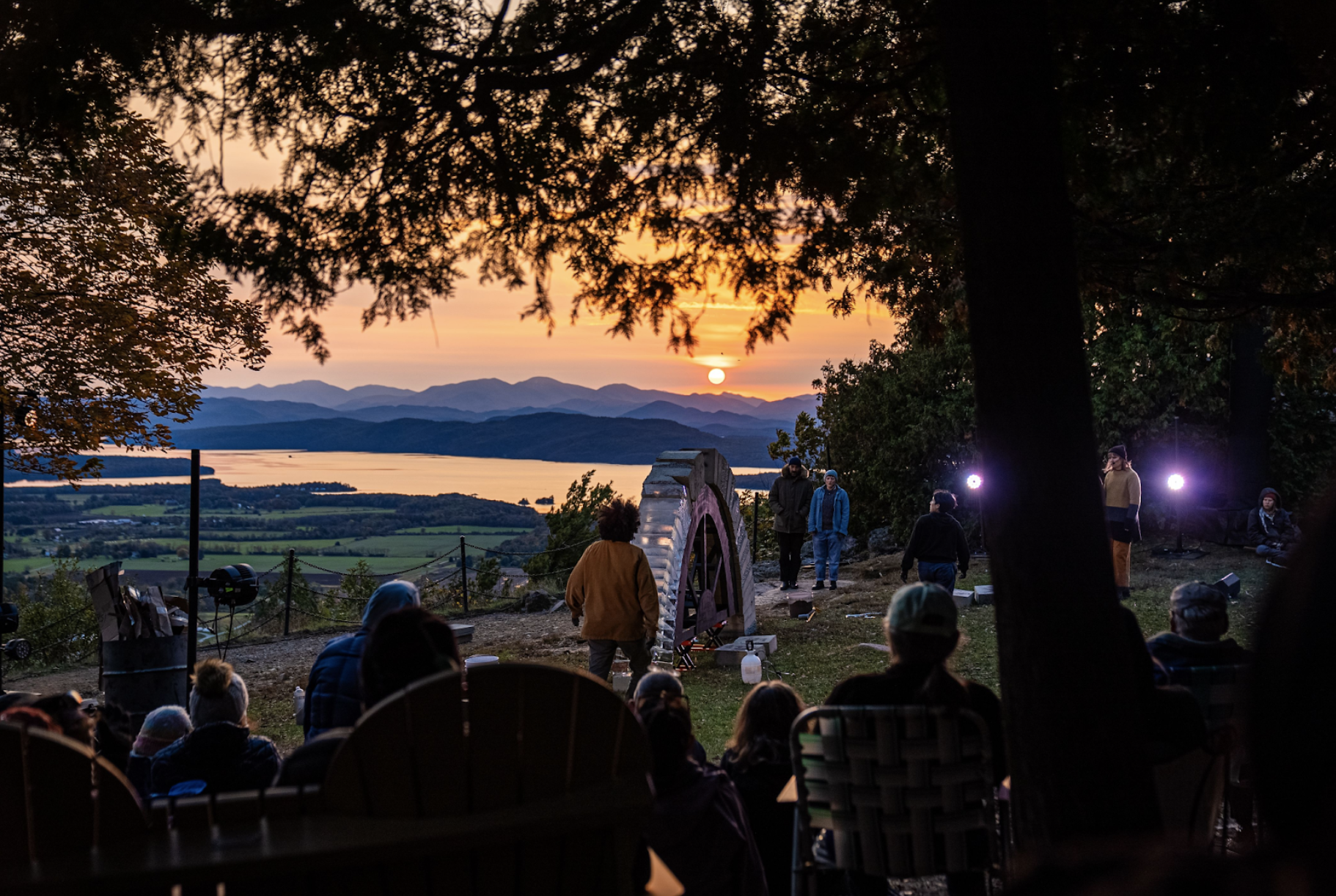 Outside. An audience and the overhanging leaves of a tree are silhouetted against the setting sun. They are watching 6 performers who are standing around a completed, freestanding arch made two thirds of concrete blocks and one third of ice blocks. Beyond, the round, orange sun is setting over a blue mountain range. at the base of the mountains is a lake, which reflects the light of the sky. 